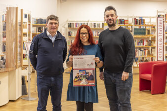 drei Personen stehen in einer Bibliothek, eine hält ein Plakat in der Hand