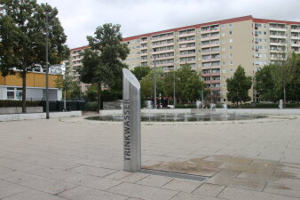 Ein stählerner Trinkbrunnen als Säule auf einer gepflasterten Fläche neben einem Springbrunnen.