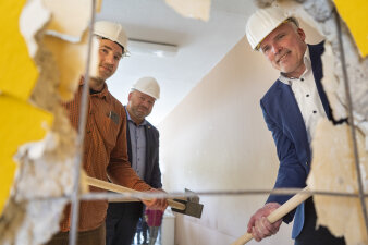 Drei Männer mit Bauhelm auf dem Kopf und Vorschlaghammer in der Hand schauen durch ein Loch in einer Wand. 