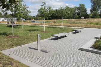 Ein stählerner Trinkbrunnen als Säule auf einer gepflasterten Fläche in einem Park. 