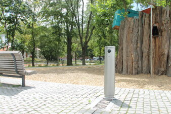 Ein stählerner Trinkbrunnen als Säule auf einer gepflasterten Fläche in einem Park. 