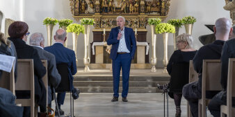 Ein Mann mit Mikrofon in der Hand steht vor einem Publikum und spricht. 