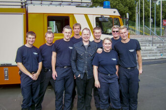 Acht Männer und Frauen in Feuerwehruniform posieren. In der Mitte steht ein Mann in Zivilkleidung. 