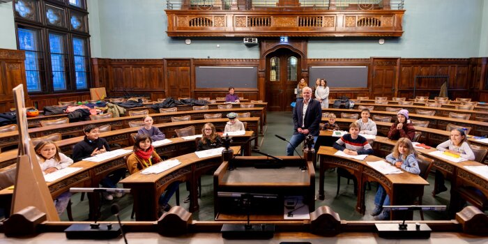 Vier Erwachsene und 14 Kinder sind zusammen in einem historischen Sitzungssaal.