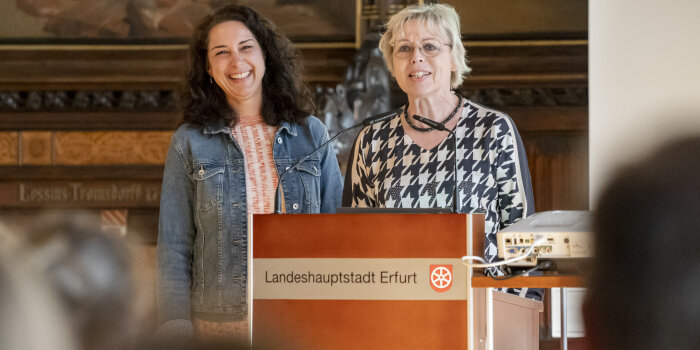 Zwei Frauen stehen an einem Rednerpult und lächeln den Besuchern im Saal zu. 