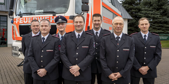 Sieben Männer in Feuerwehruniform posieren vor einem Feuerwehrfahrzeug.