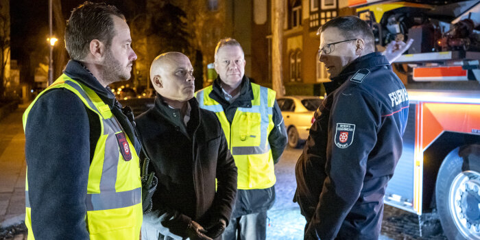 Vier Männer stehen im Dunkeln vor einem Feuerwehrfahrzeug und unterhalten sich. Zwei davon tragen eine Warnweste. 