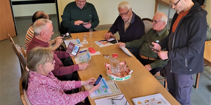 Sechs ältere Menschen üben den Umgang mit dem Smartphone. Ein Mann steht dabei und zeigt den Umgang.