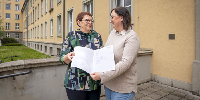 Zwei Frauen stehen beisammen und halten gemeinsam eine Urkunde in der Hand. 