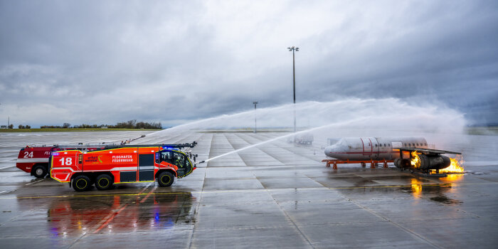 Löschfahrzeuge der Feuerwehr spritzen Wasser auf einen brennenden Flugzeugrumpf. 