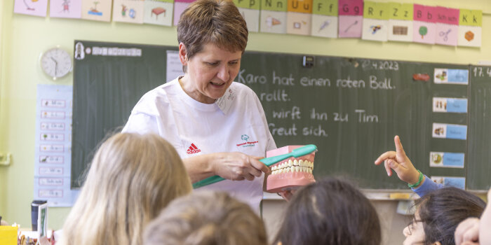 Eine Frau demonstriert Kindern an einem übergroßen Zahnmodell das richtige Zähneputzen.