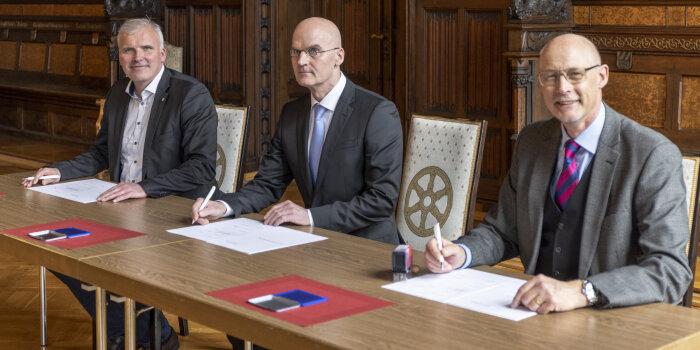 Drei Männer sitzen an einem Tisch und unterzeichnen einen Vertrag.