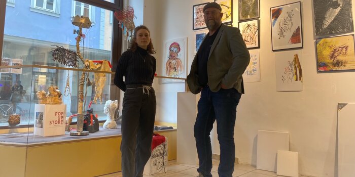 Zwei Personen stehen in einer Galerie mit den ausgestellten Kunstwerken im Hintergrund. 