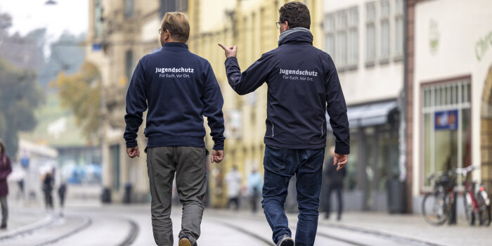 Rückansicht zweier Männer, die in Dienstkleidung durch eine Straße laufen und gestikulieren.