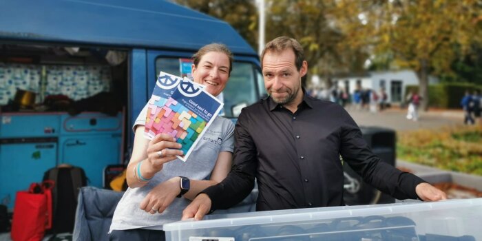 Eine Frau und ein Mann. Die Frau zeigt Flyer und der Mann trägt eine Plastikkiste.