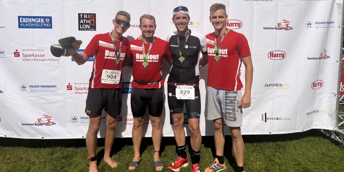 Vier Männer in Sportkleidung stehen jubelnd vor einem Werbeplakat.