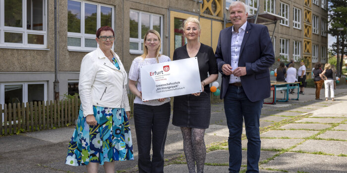 Drei Frauen und ein Mann präsentieren ein Schild, auf dem der Name einer Schule steht.