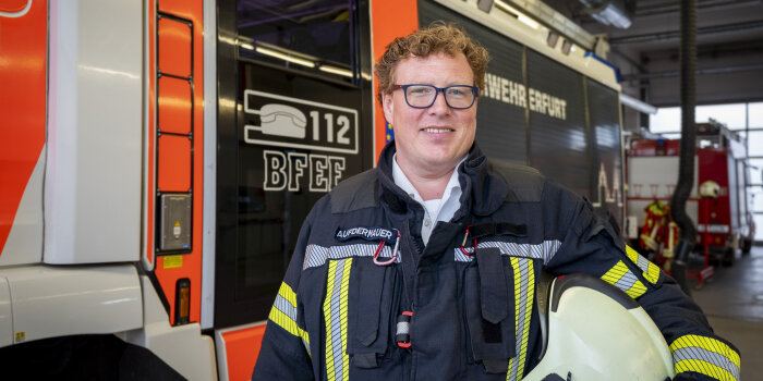 Ein Mann mit Brille und lockigen Haaren trägt Feuerwehrmontur und steht vor einem Feuerwehrfahrzeug.