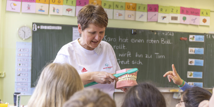 Eine Frau demonstriert Kindern an einem übergroßen Zahnmodell das richtige Zähneputzen.