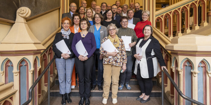 Auf einer Treppe stehen 25 Frauen und Männer und halten Mappen mit einem Vertrag in der Hand.