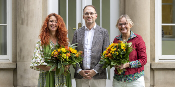 drei Personen, zwei davon mit einem Blumenstrauß