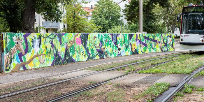 Tiere und Pflanzen als buntes Graffiti auf einer Mauer. Am rechten Bildrand ist eine Straßenbahn sichtbar.