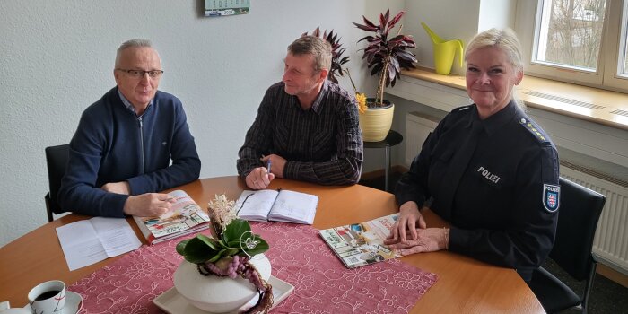 Zwei Männer in zivil und eine Frau in Polizeiuniform sitzen an einem Tisch. Die Männer unterhalten sich.