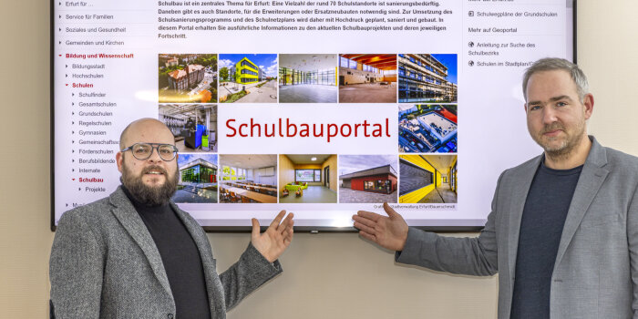 Zwei Männer stehen vor einem großen Bildschirm mit der Aufschrift Schulbauportal.