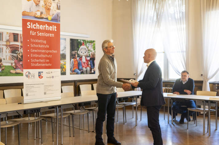 Ein Mann mit Glatze gibt einem älteren Mann einen Handschlag. Im Hintergrund sitzt ein weiterer Mann. 