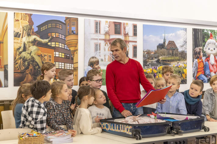 Ein Mann im roten Pullover holt eine Mappe aus einem Koffer. Um ihn herum stehen viele Kinder. 