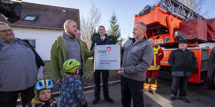 Ein Mann übergibt ein Schild an einen anderen Mann. Dabei stehen weitere Männer und zwei Kinder. 