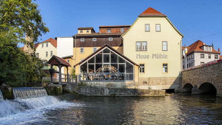 Ein Haus mit der Aufschrift „Neue Mühle“. Davor fließt Wasser in einem Fluss unter einer Brücke durch.