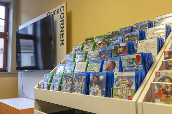 In einem Regal stehen viele Videospiele. Links daneben hängt ein Bildschirm. Daran steht: Game Corner. 