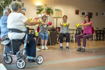 Fünf Frauen und ein Mann sitzen im Stuhlkreis und halten einen Ball. Eine Teilnehmerin hat einen Rollator.