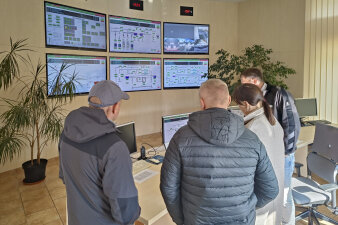 vier Personen stehen in einer Schaltzentrale mit sechs Monitoren an der Wand