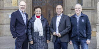 Drei Männer und eine Frau, alle dunkelblau gekleidet, stehen nebeneinander. 