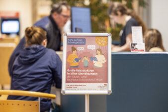 Ein Schild weist auf eine Rabattaktion in einer Bibliothek hin. Im Hintergrund sind Menschen im Gespräch. 