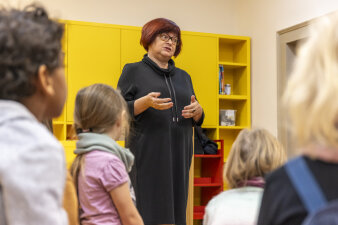 Eine ältere Frau mit rötlichen Haaren steht vor Kindern und spricht. 