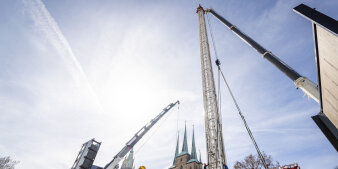 Der Turm eines Kettenkarussells wird aufgebaut mit Kranen. Im Hintergrund ist ein Dom zu sehen.