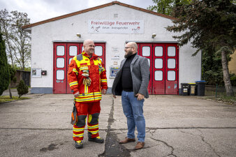 Vor einem Feuerwehrgerätehaus stehen zwei Männer. Einer in Feurwehruniform, der andere in Jackett und Jeanshose. 