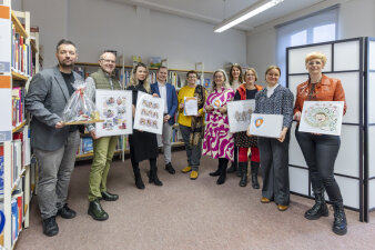 Zehn Männer und Frauen stehen in einem Raum mit Büchern. Sie halten Bilder, Geschenke und Zettel in der Hand.