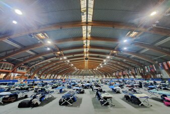 In einer großen Sporthalle stehen hunderte aufgebaute Feldbetten. 