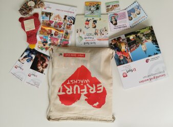 Flyer, ein Turnbeutel mit Aufschrift und Anziehsachen für Kinder liegen auf einem Tisch.