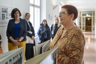 Eine Frau mit kurzen roten Haaren und Brille lächelt an einem Pult stehend. Im Hintergrund stehen weitere Menschen.