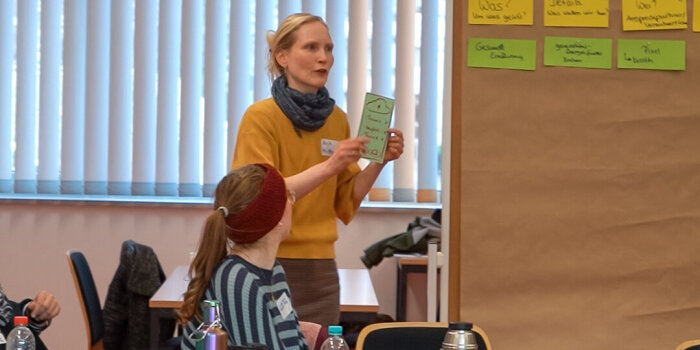 Eine blonde Frau im gelben Pullover spricht vor anderen Menschen. Sie hält dabei eine grüne Karte in der Hand.