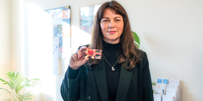 Eine Frau mit langen brünetten Haaren trägt einen grünen Blazer und schwarzen Pullover. Sie zeigt eine Visitenkarte