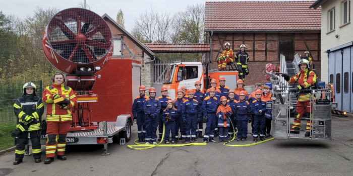 Sechs Erwachsene und zahlreiche Kinder in Feuerwehrkleidung posieren zum Gruppenfoto an Feuerwehrgerätschaften 