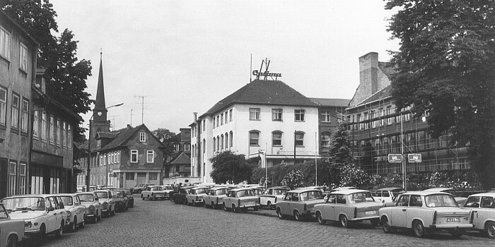 an einer Straße stehen viele Trabanten, im Mittelpunkt ein Gebäude mit Schriftzug Optima