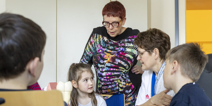 Eine Frau sitzt mit Kindern an einem Tisch und erklärt etwas. Dabei steht eine weitere Frau und sieht zu. 