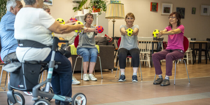 Fünf Frauen und ein Mann sitzen im Stuhlkreis und halten einen Ball. Eine Teilnehmerin hat einen Rollator.